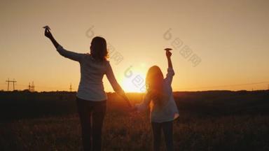 妈妈和女儿在日落时正在玩纸飞机。在风景如画的地方。快乐的时间与婴儿的概念